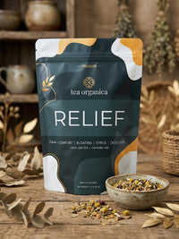 A stand-up pouch of tea organica Relief loose leaf tea is displayed on a rustic wooden table with a small bowl of the loose leaf blend and scattered dried leaves in the foreground. The background is a warm, softly blurred wooden shelf setting.