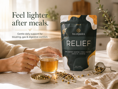 A stand-up pouch of tea organica Relief tea is shown on a tabletop beside a glass cup of amber tea, loose tea pieces, and a small metal infuser. A person's hands are visible holding the cup, and the scene is styled with a vase and plants in a softly lit interior.